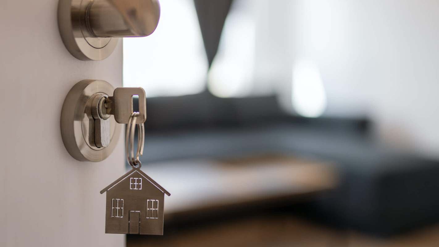 A key with a house-shaped keychain hangs from a door lock, with a blurred room visible in the background.