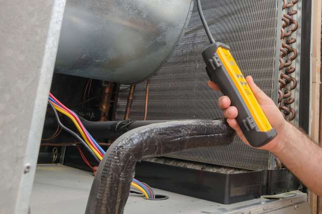 A person holds a diagnostic tool while interacting with an air conditioning unit's internal components.