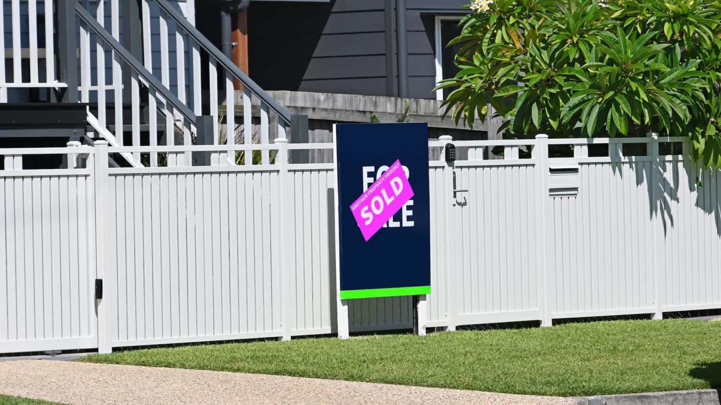 A "Sold" sign on a fence in front of a property with a house and greenery.