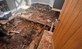 A room under renovation with exposed flooring, scattered debris, and a partially removed section of the floor.