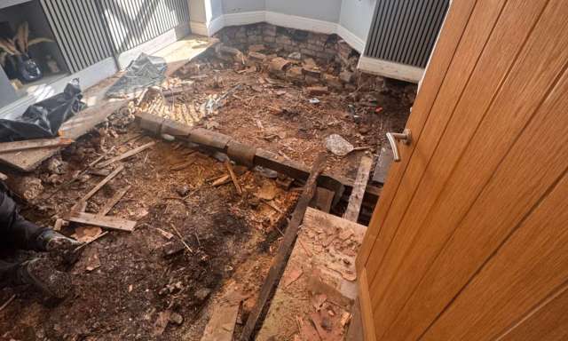 A room under renovation with exposed flooring, scattered debris, and a partially removed section of the floor.