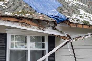A damaged roof covered with a blue tarp showing exposed wood and debris near a window.