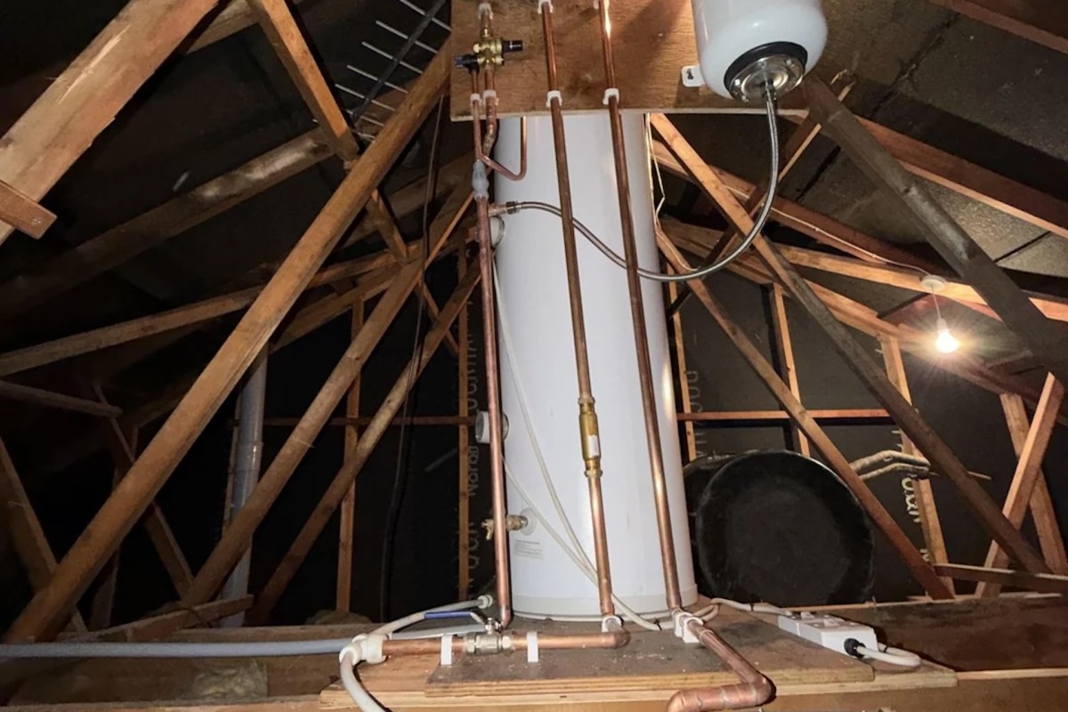 A water heater and plumbing pipes in an attic with exposed wooden beams.