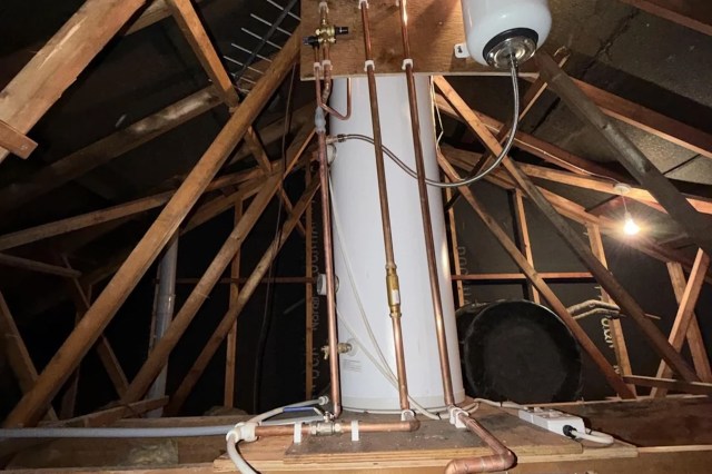 A water heater and plumbing pipes in an attic with exposed wooden beams.