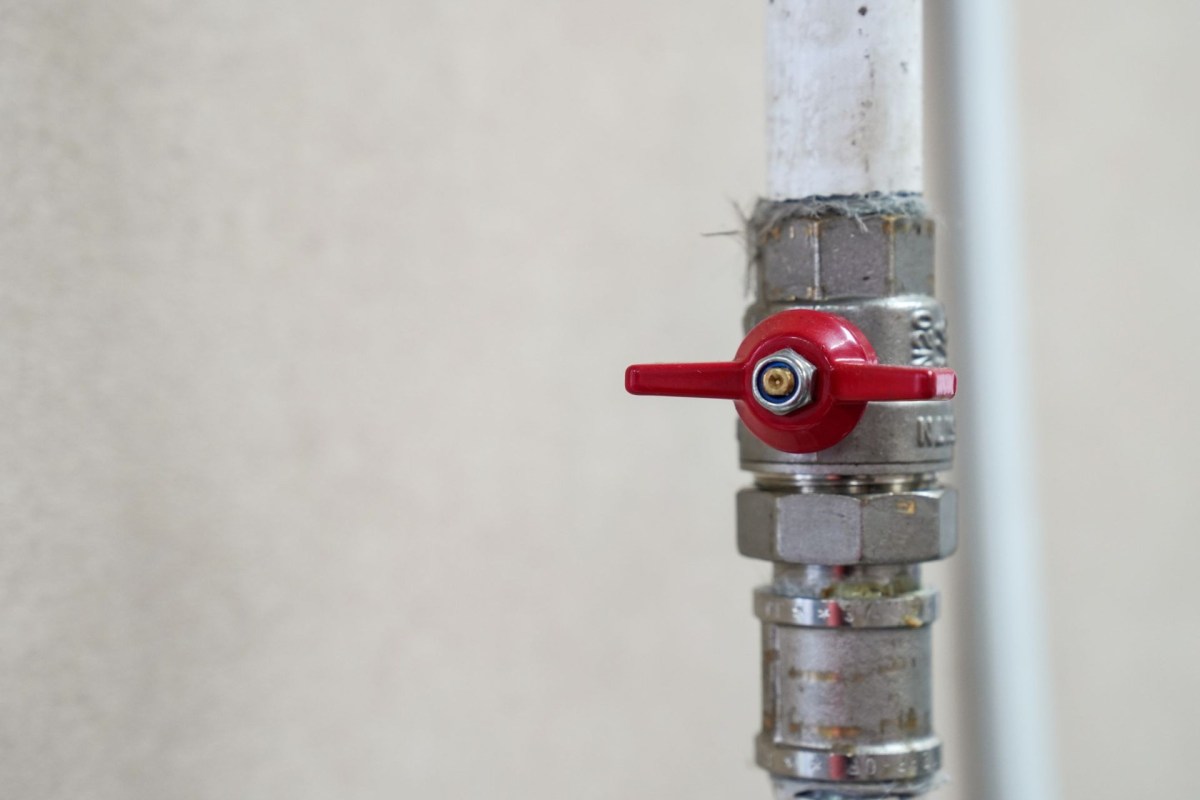 A red water shut off valve attached to a metal pipe against a light-colored wall.
