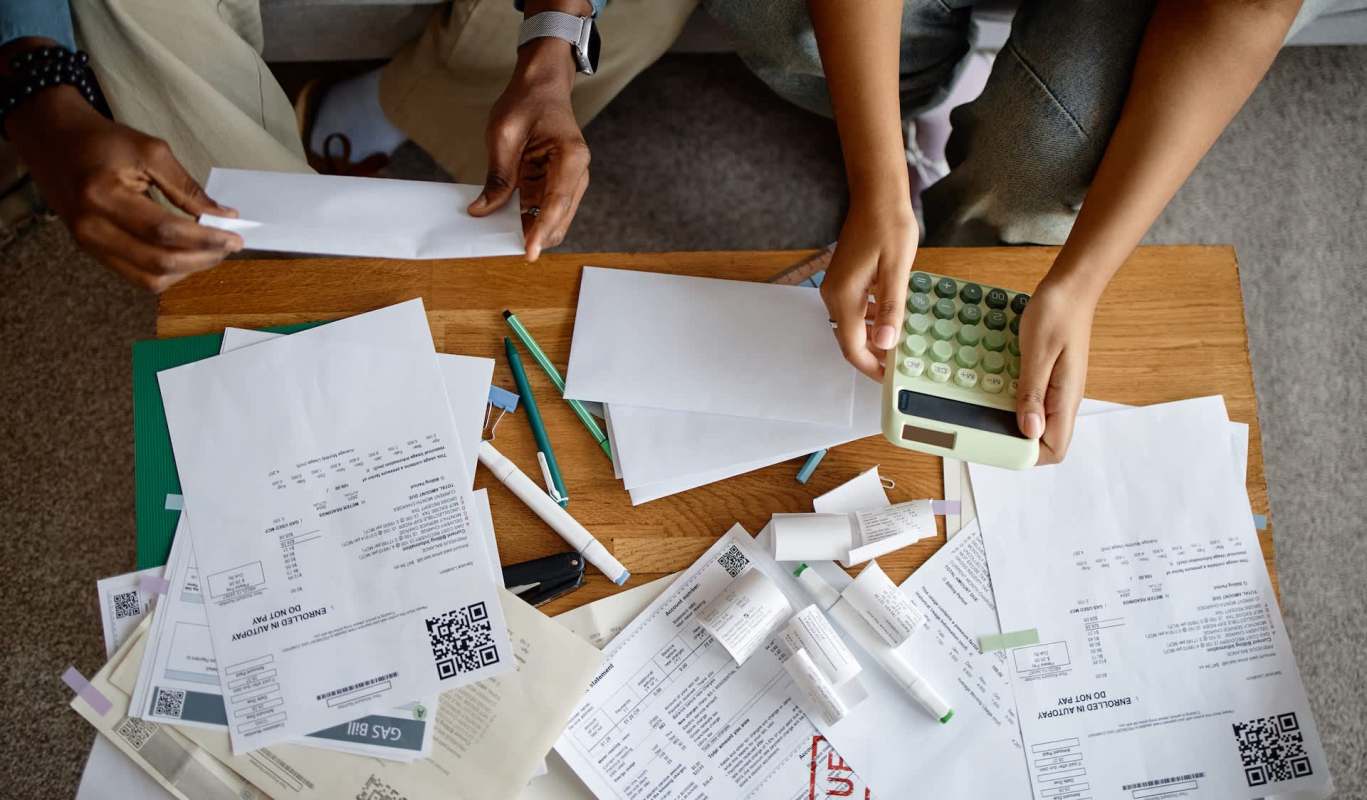Two people handle paperwork and a calculator on a wooden table covered with various documents and stationery.