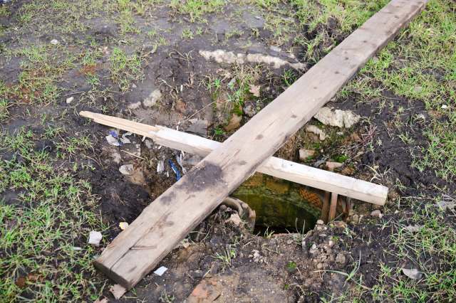 A hole in the ground covered with wooden planks, surrounded by grass and debris.