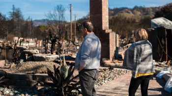 Two people stand amid the ruins of a burned landscape, observing the destruction around them.