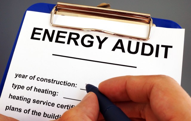 A hand holds a pen above a clipboard titled "Energy Audit" with blank fields for information.