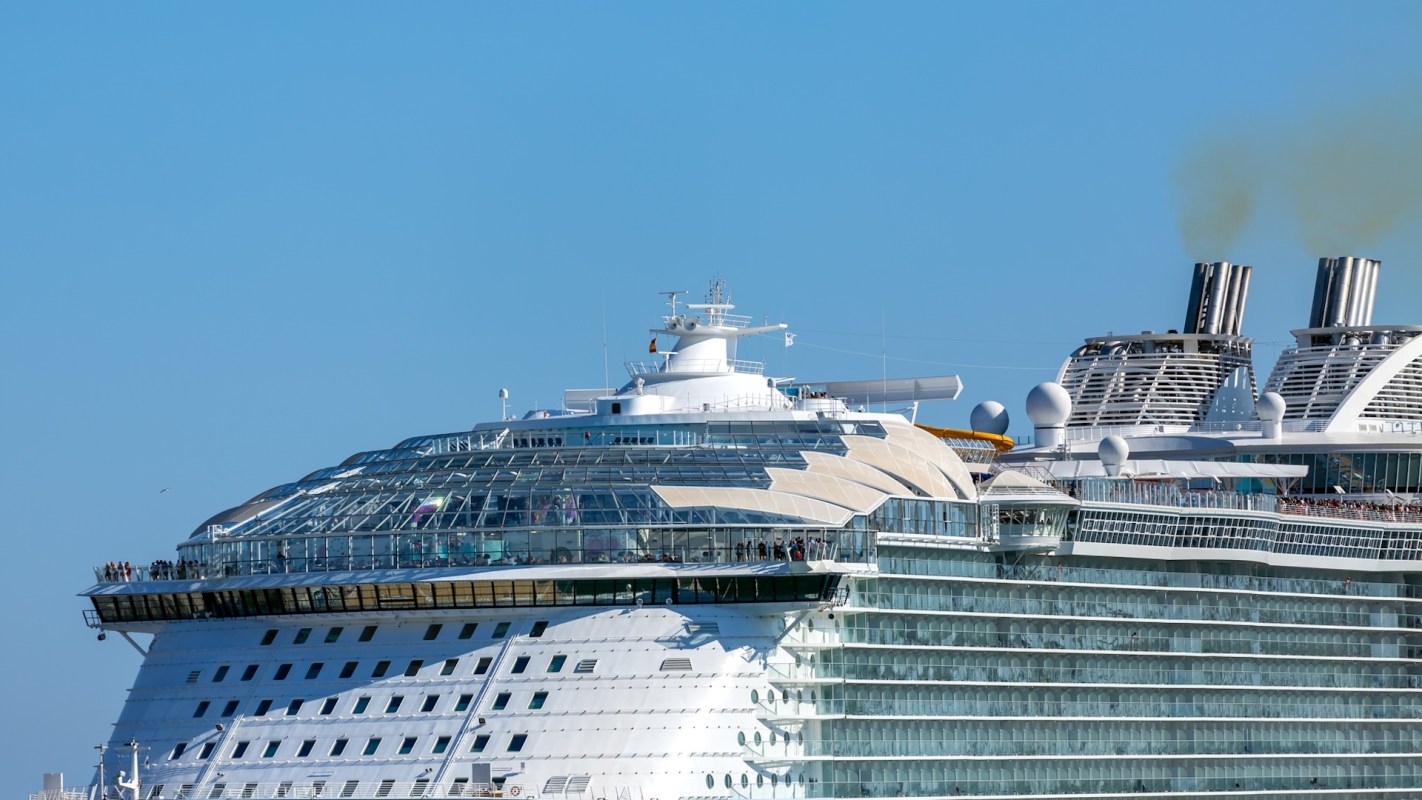 A large cruise ship with a panoramic glass roof and smoke stacks against a clear blue sky.