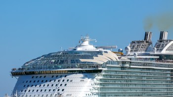 A large cruise ship with a panoramic glass roof and smoke stacks against a clear blue sky.