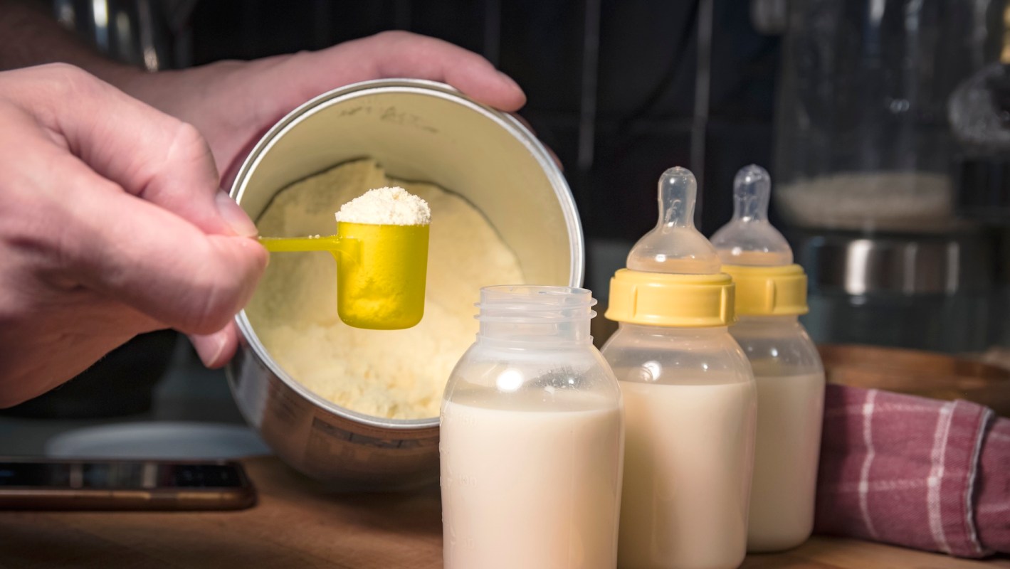 A hand holds a yellow scoop filled with powder near baby bottles filled with liquid.