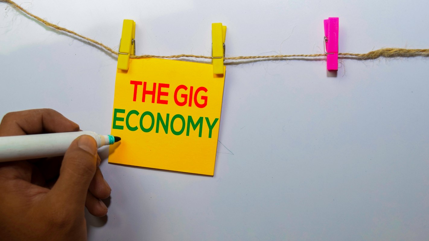 A hand holds a marker near a yellow sticky note that says "THE GIG ECONOMY," hanging on a string with clothespins.