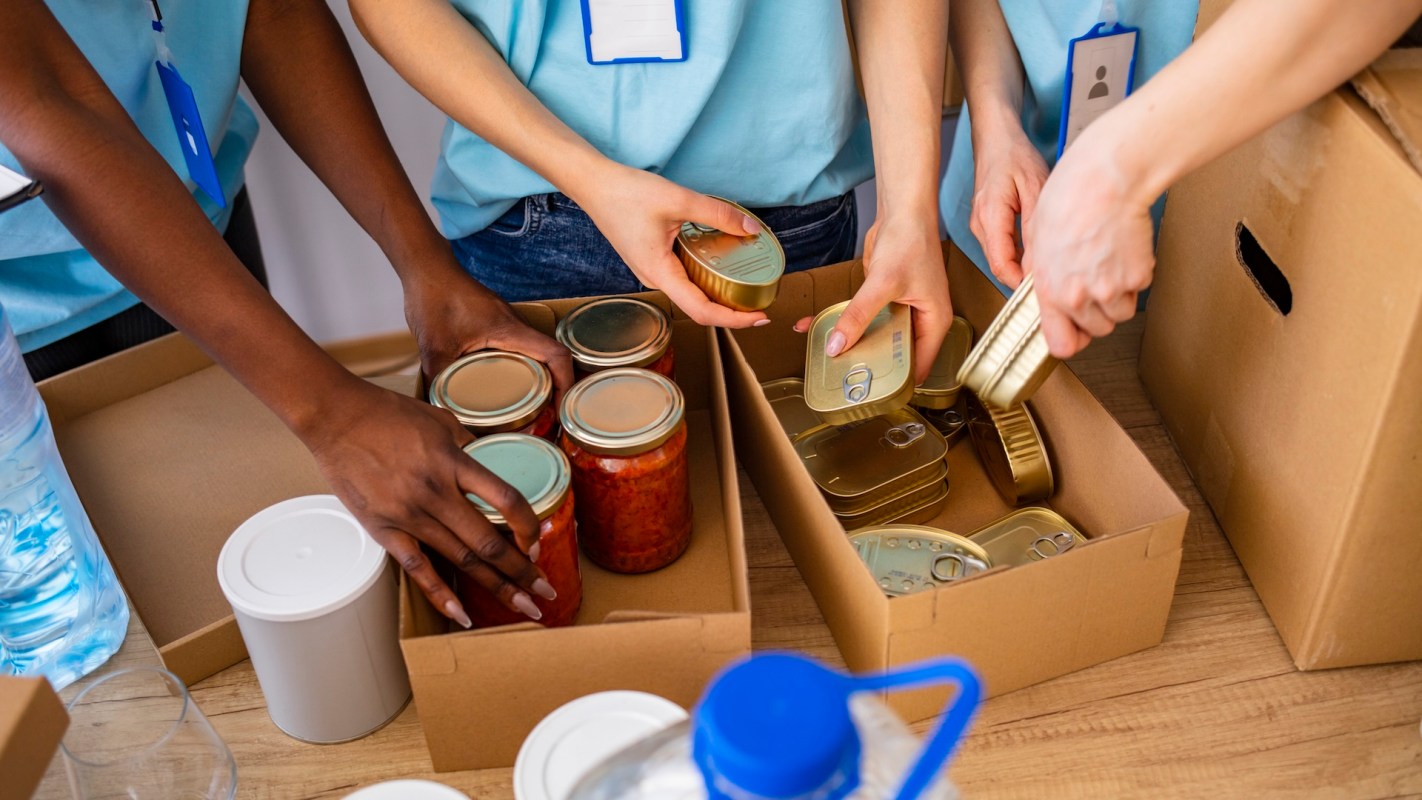 People organize canned and jarred food items into boxes on a table.