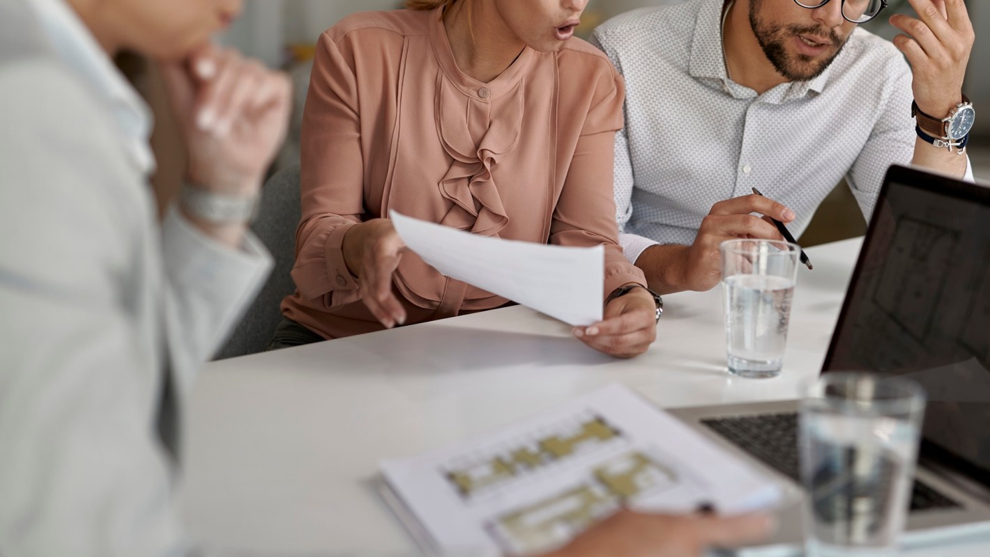 A man and woman looking displeased at documents with a real estate agent holding floor plans of a building.
