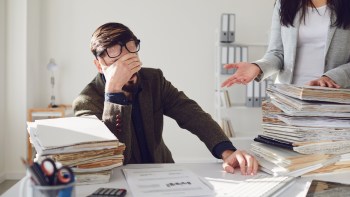 A man sits at a desk with his head in his hand, while a woman gestures with papers stacked nearby.