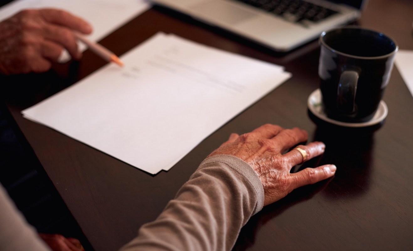 An elderly woman's hand rests on a table with papers and a coffee cup nearby, while someone prompts her to sign a paper.