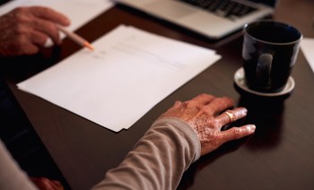 An elderly woman's hand rests on a table with papers and a coffee cup nearby, while someone prompts her to sign a paper.