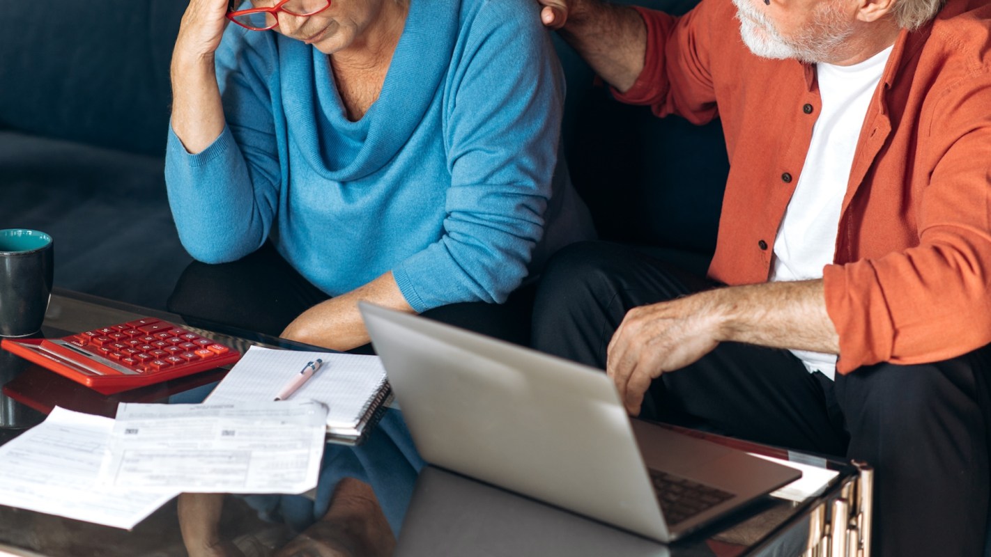 An older woman sits at a table, looking down, with papers and a laptop while a man gestures toward her.
