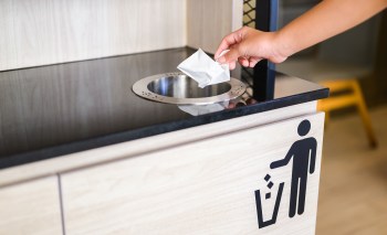 A hand drops a piece of paper into a trash bin on a wooden cabinet.