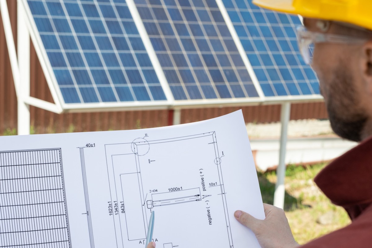 A person holds a blueprint while examining solar panels in the background.