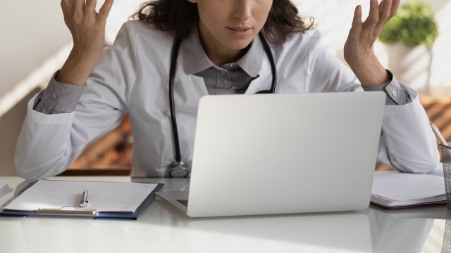 A person in a white coat gestures while looking at a laptop on a desk with papers and a pen.