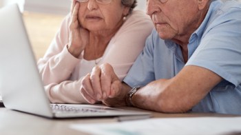 An elderly couple is looking at a laptop together while sitting at a table.