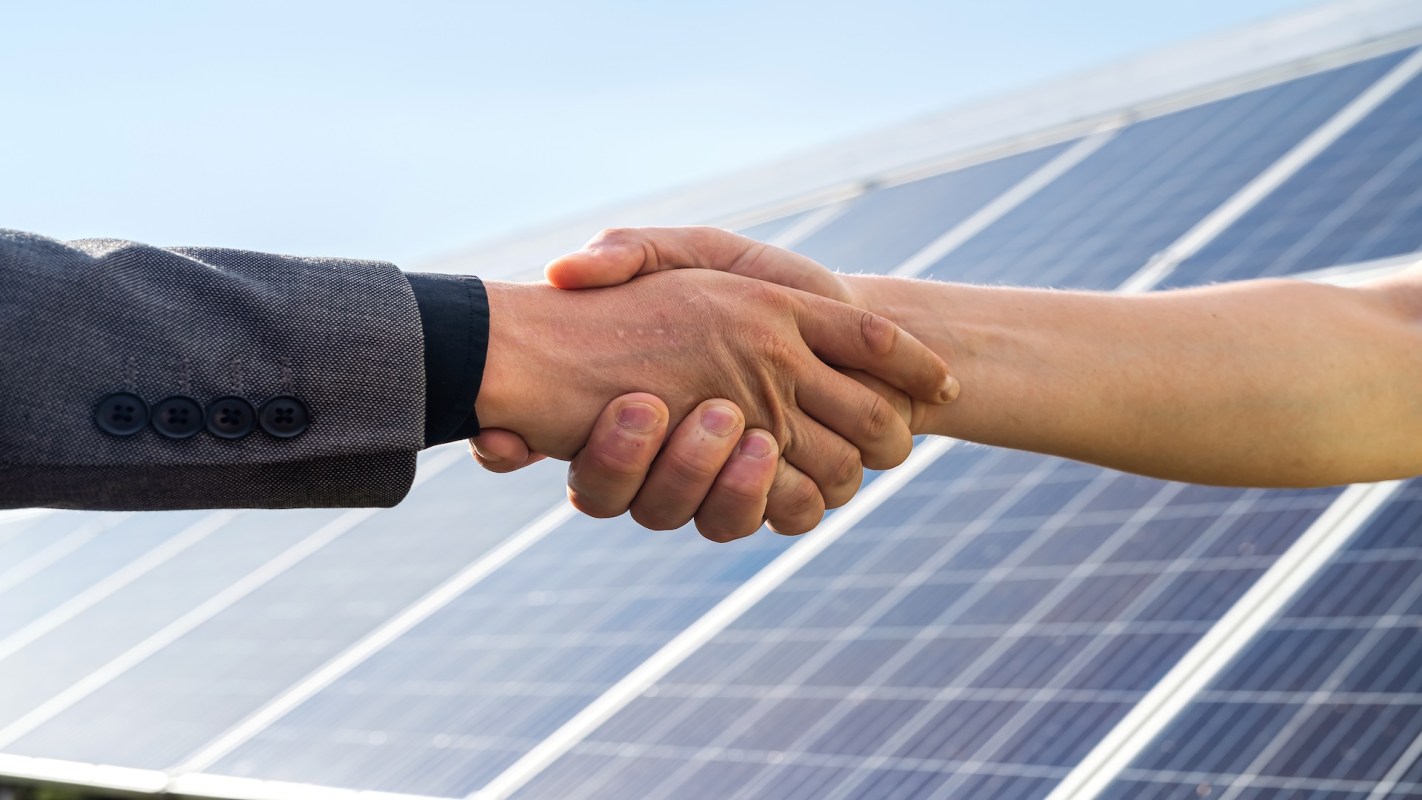 Two hands shake in front of solar panels.