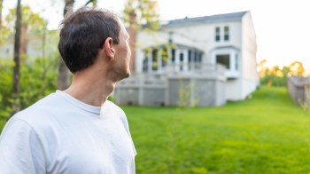 A man stands in a green yard, looking toward a house in the distance.