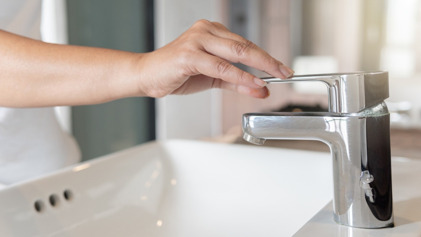 A hand adjusts a faucet above a white sink in a modern bathroom.