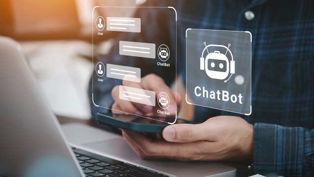 A person using a smartphone with a laptop in front, displaying chatbot conversation interfaces.