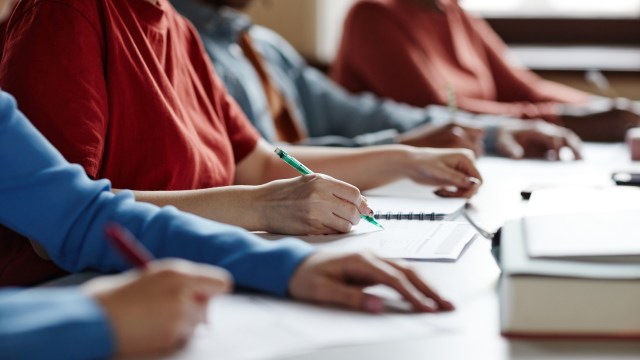 Several students are writing in notebooks at a table with papers and books.