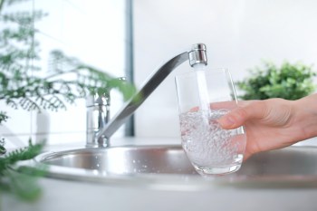 A hand fills a glass with water from a faucet in a kitchen sink, with plants in the background.