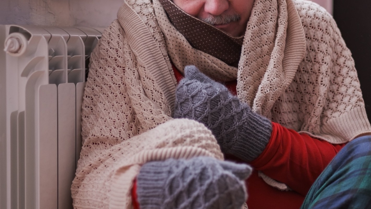 A person wrapped in blankets sits next to a radiator wearing mittens.