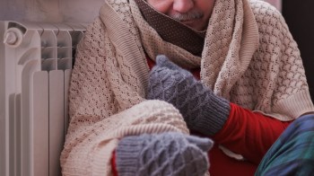 A person wrapped in blankets sits next to a radiator wearing mittens.
