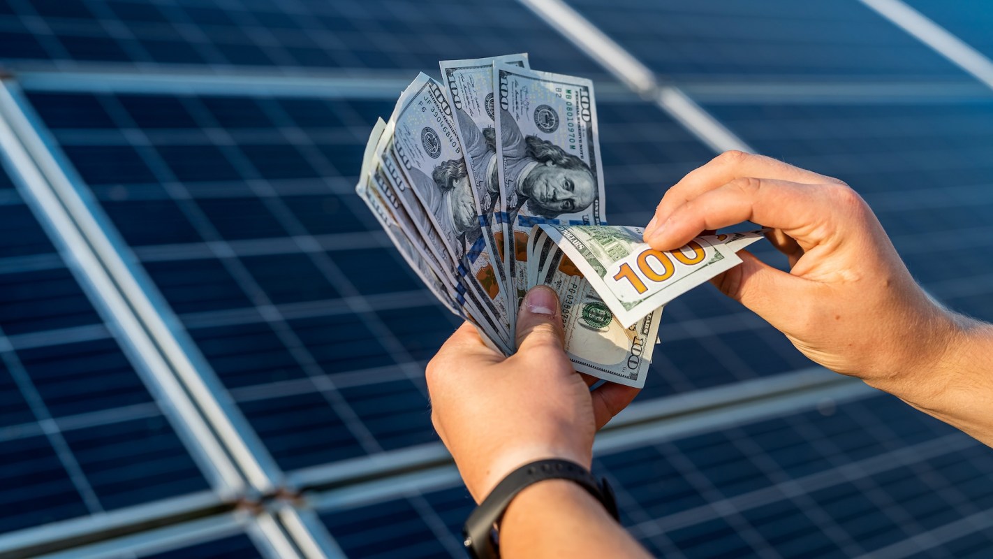 Hands hold a fan of hundred-dollar bills above solar panels.