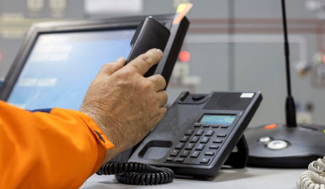 A person holds a phone near a desk with a computer and a landline phone.