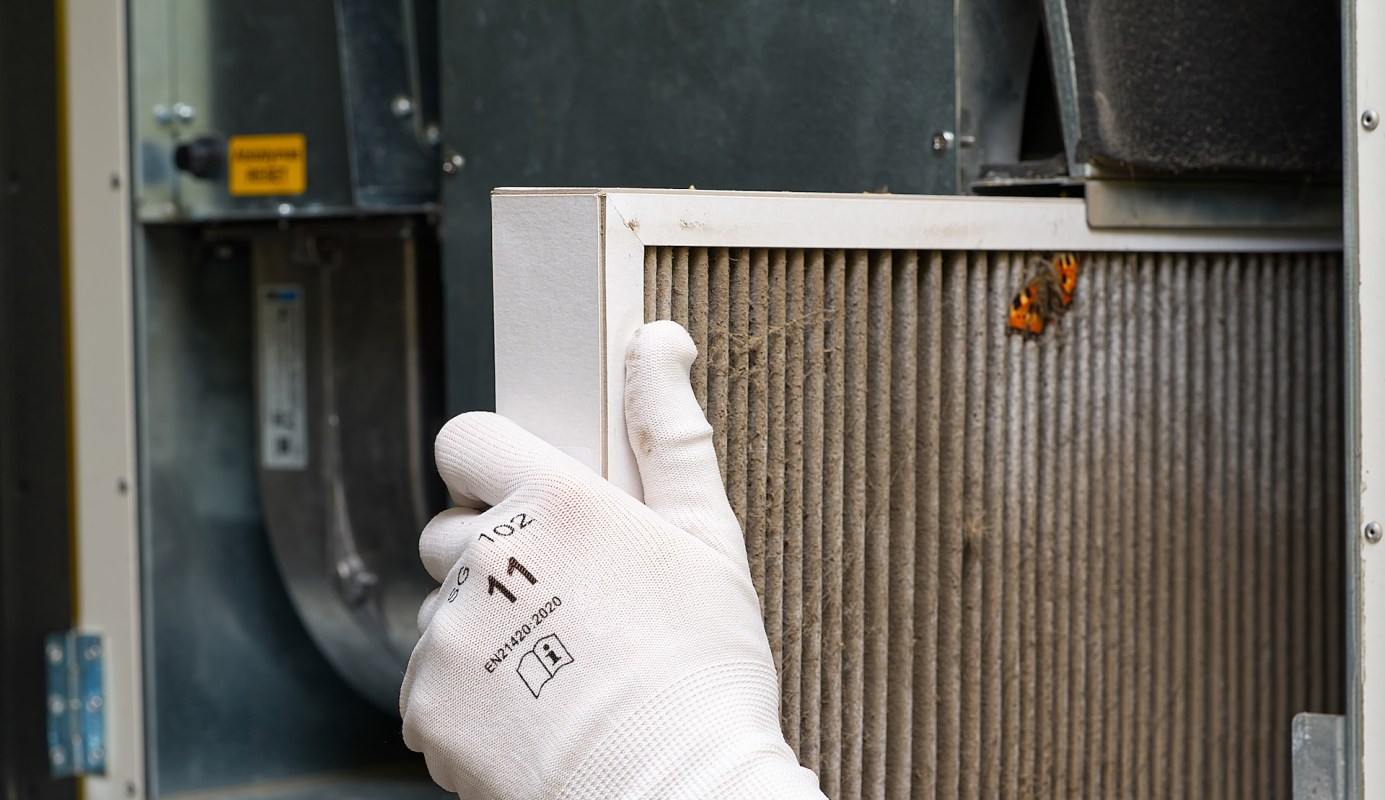 A person wearing gloves removes a dirty air filter from an HVAC unit.