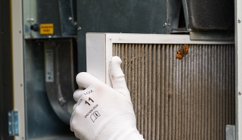 A person wearing gloves removes a dirty air filter from an HVAC unit.