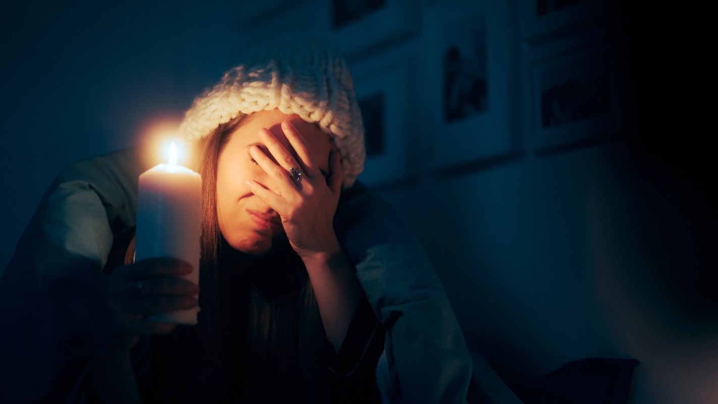 A woman sitting in bed holds a candle in the dark while covering her face with her other hand.