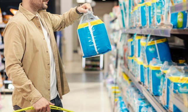 A person holding a blue package of diapers in a store aisle with shelves of products in the background.