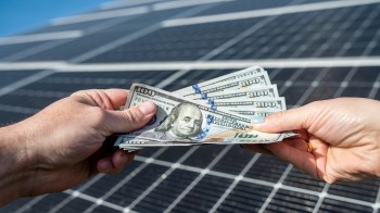 Two hands exchange several hundred dollar bills in front of a solar panel.