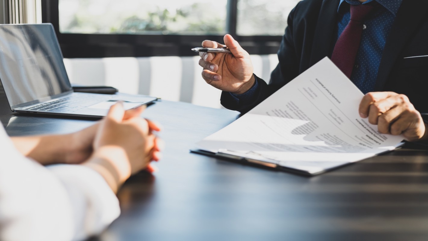 A person holds a pen and documents while another person sits at a desk with hands clasped.