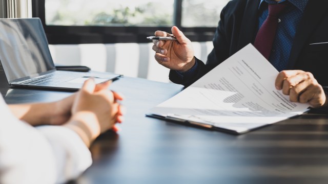 A person holds a pen and documents while another person sits at a desk with hands clasped.