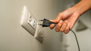 A hand holds a plug near a wall electrical outlet.