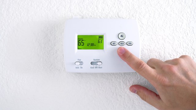 A hand adjusts the temperature on a digital thermostat displaying indoor and hold settings.