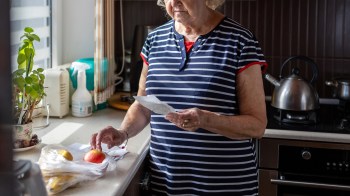 A senior woman looks at a receipt with concern, with some fruit on a kitchen counter.