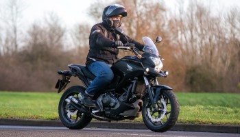 A person riding a black electric motorcycle on a road beside grass and trees.