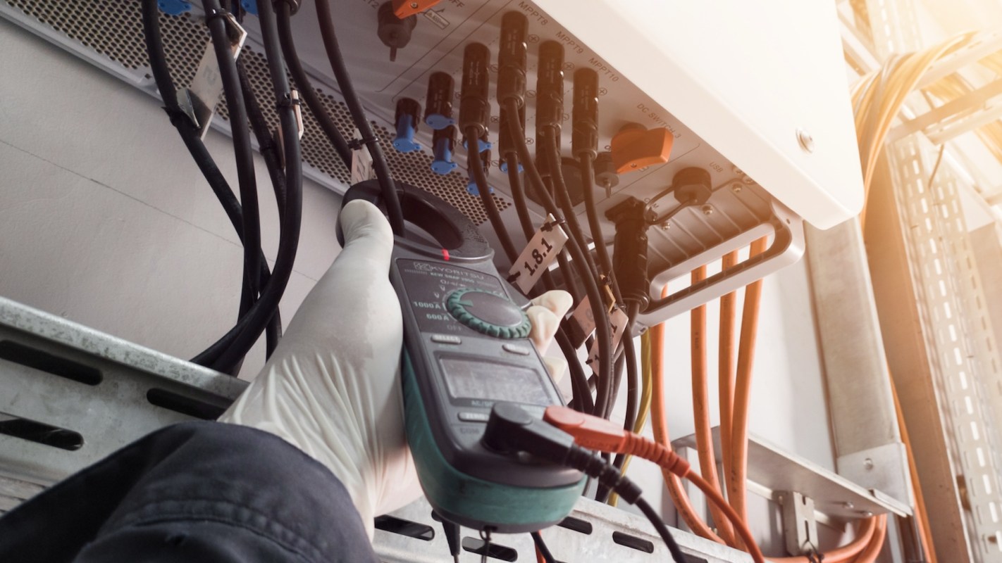 A person holds a multimeter while checking connections on a solar inverter with various colored wires.