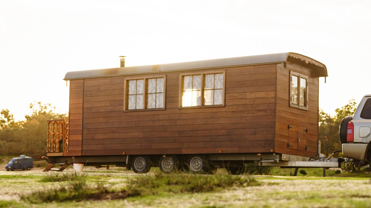 A wooden trailer home parked on grass with a vehicle attached.